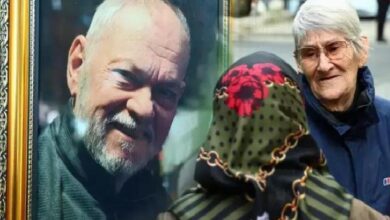 An elderly couple on a street examines a framed portrait; the glass reflects a smiling man’s face.