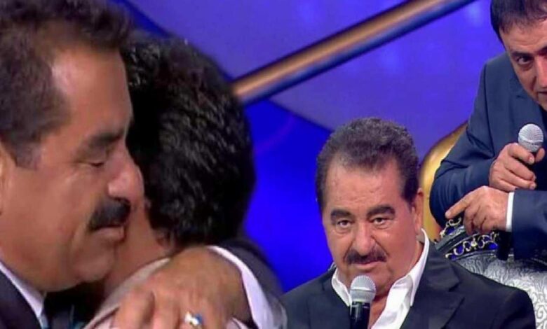 Close-up of a man in a dark suit speaking into a handheld microphone on a blue-lit stage, with another man in the foreground and a third man in the background holding a microphone.