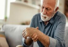Elderly man with a white beard sits on a sofa, looking down at his wrist as if in pain or discomfort.
