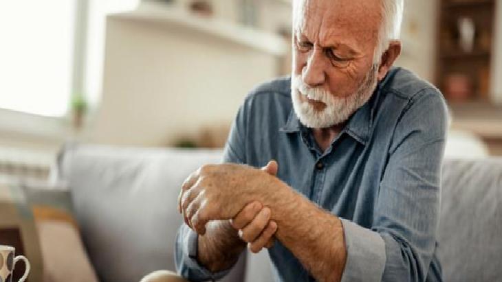 Elderly man with a white beard sits on a sofa, looking down at his wrist as if in pain or discomfort.