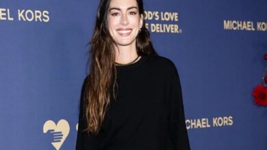 Smiling woman with long wavy hair in a black top posing against a blue Michael Kors backdrop with a heart logo on the left.
