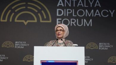 Woman wearing a light hijab and patterned coat speaks at a podium with microphones at the Antalya Diplomacy Forum backdrop.