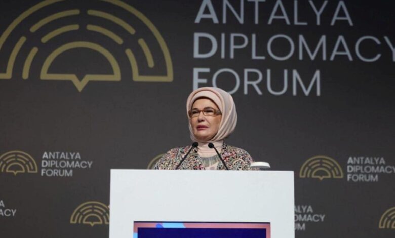Woman wearing a light hijab and patterned coat speaks at a podium with microphones at the Antalya Diplomacy Forum backdrop.