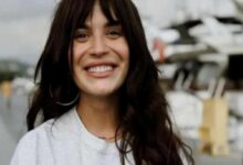 Smiling woman with long dark hair in a light gray shirt, outdoors near boats and yachts in a marina environment.
