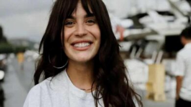 Smiling woman with long dark hair in a light gray shirt, outdoors near boats and yachts in a marina environment.
