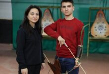 Woman and a young man stand in an indoor archery range; the man is drawing a bow while wearing a red shirt, with target boards in the background to the right and left.