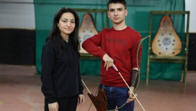 Woman and a young man stand in an indoor archery range; the man is drawing a bow while wearing a red shirt, with target boards in the background to the right and left.