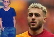 Close-up of a man with blond curls in a red shirt, looking thoughtful, with a boy in a dark blue shirt beside a water body in the background.