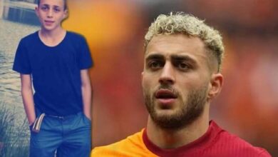 Close-up of a man with blond curls in a red shirt, looking thoughtful, with a boy in a dark blue shirt beside a water body in the background.
