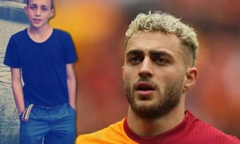 Close-up of a man with blond curls in a red shirt, looking thoughtful, with a boy in a dark blue shirt beside a water body in the background.