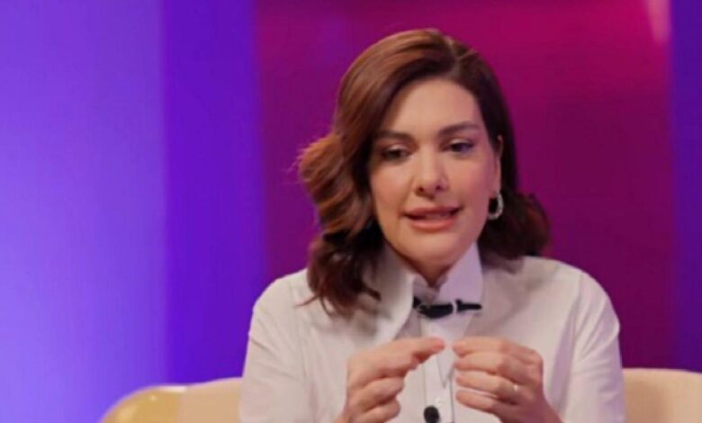 Woman in a white blouse sits on a purple-lit stage, speaking and gesturing with her hands.