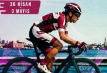 Road cyclist in pink/white kit riding along a waterfront with an event banner reading '26 Nisan 3 Mayıs' in the top left.
