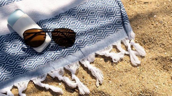 Sunglasses and a sunscreen bottle rest on a blue geometric towel spread on sandy beach.