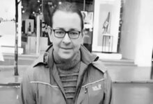Man with glasses wearing a striped turtleneck and a jacket, facing the camera on a city sidewalk with storefronts behind him (black-and-white photo).