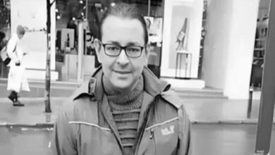 Man with glasses wearing a striped turtleneck and a jacket, facing the camera on a city sidewalk with storefronts behind him (black-and-white photo).
