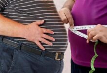A person in a striped shirt holds their stomach while another person uses a measuring tape around their waist to take a measurement.