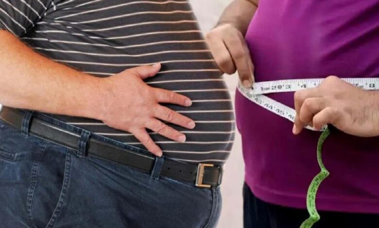 A person in a striped shirt holds their stomach while another person uses a measuring tape around their waist to take a measurement.