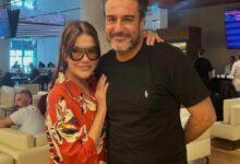 Couple posing for a photo in a modern lounge; woman in a red floral blouse and sunglasses, man in a black t-shirt, arms around each other, blue-lit background with digital boards.