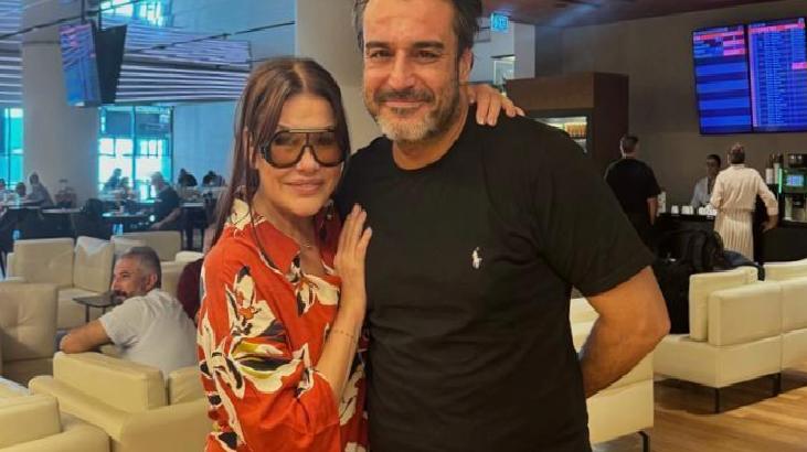 Couple posing for a photo in a modern lounge; woman in a red floral blouse and sunglasses, man in a black t-shirt, arms around each other, blue-lit background with digital boards.