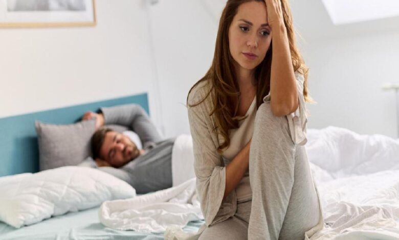 Upset woman sitting on the edge of a bed, hand on her head, while a man sleeps in the background.