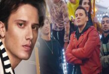 Close-up of a young man in the foreground with a striped scarf, others posing behind in a brightly lit hallway or studio.