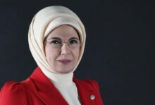 Professional portrait of a woman wearing a beige hijab, glasses, and a red blazer against a dark background.