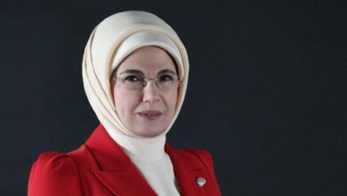 Professional portrait of a woman wearing a beige hijab, glasses, and a red blazer against a dark background.