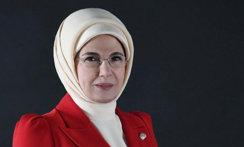 Professional portrait of a woman wearing a beige hijab, glasses, and a red blazer against a dark background.