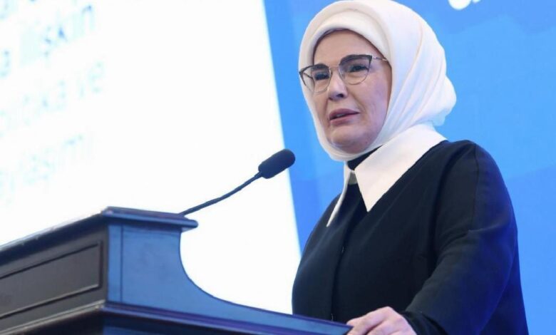 Woman wearing a white hijab and glasses speaks at a black podium with a microphone at a conference booth/stage, blue backdrop behind her.