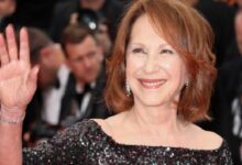 Smiling woman in a sequined off-shoulder dress waves to the camera on a red carpet.