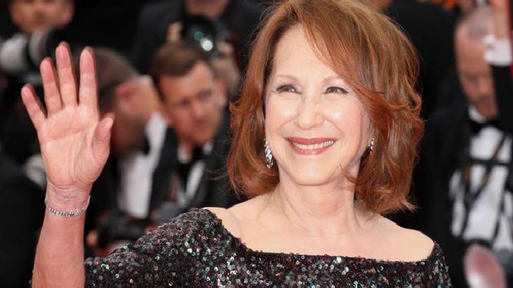 Smiling woman in a sequined off-shoulder dress waves to the camera on a red carpet.