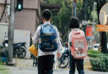Two children with backpacks walking down a city sidewalk, blue backpack on the left and pink cat-faced backpack on the right.