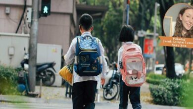 Two children with backpacks walking down a city sidewalk, blue backpack on the left and pink cat-faced backpack on the right.