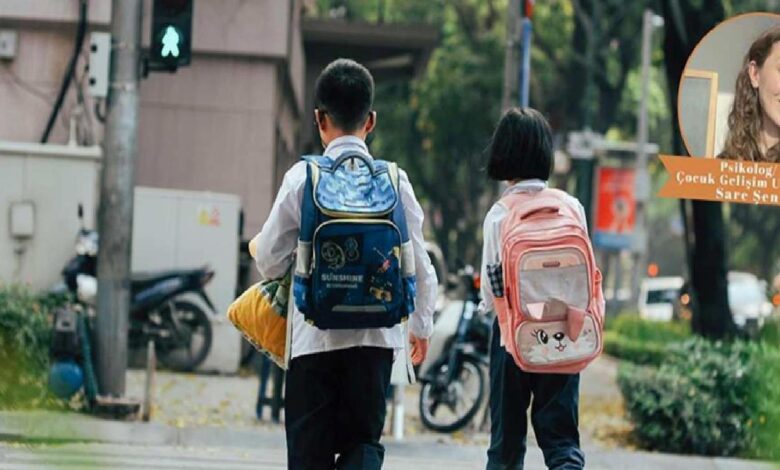 Two children with backpacks walking down a city sidewalk, blue backpack on the left and pink cat-faced backpack on the right.