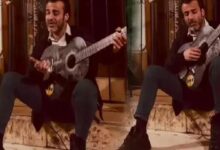 Man with a beard sits on marble steps indoors, playing a gray acoustic guitar.