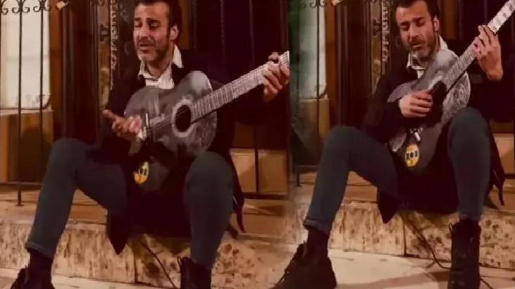 Man with a beard sits on marble steps indoors, playing a gray acoustic guitar.