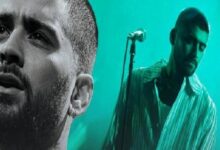 Two male performers on a live stage under teal lighting, one in the foreground close-up and the other near a microphone in the background.