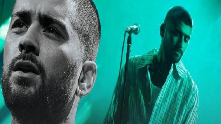 Two male performers on a live stage under teal lighting, one in the foreground close-up and the other near a microphone in the background.