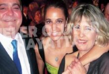 Three adults in formal attire posing together at a crowded social event, with a man in a suit and blue tie on the left and two women embracing in the center and right.