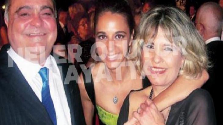 Three adults in formal attire posing together at a crowded social event, with a man in a suit and blue tie on the left and two women embracing in the center and right.