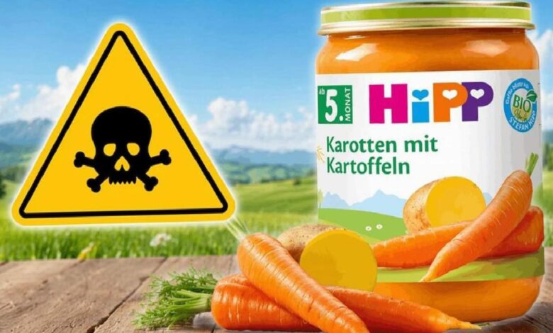 HiPP carrot-and-potato baby food jar on a wooden surface with fresh carrots, and a yellow skull-and-crossbones warning sign in a countryside background.