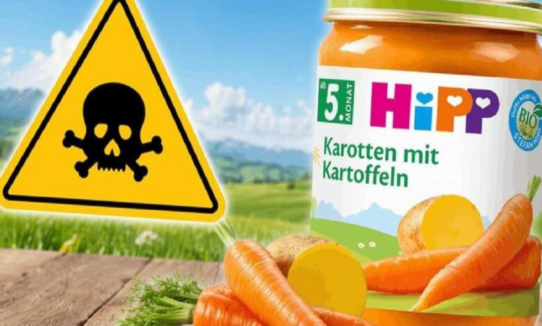 Yellow caution triangle with skull and crossbones beside a HiPP carrot-and-potato baby food jar in a rural outdoor setting.