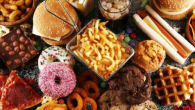 Assorted fast foods and desserts on a table: burgers, fries, donuts, pizza slice, waffles, hot dogs, chocolate bar and more jam-packed together.