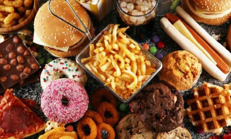 Assorted fast foods and desserts on a table: burgers, fries, donuts, pizza slice, waffles, hot dogs, chocolate bar and more jam-packed together.