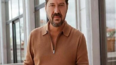 Man with short dark hair and beard wearing a tan zip-neck sweater stands in a glass-walled corridor or atrium, looking toward the camera.