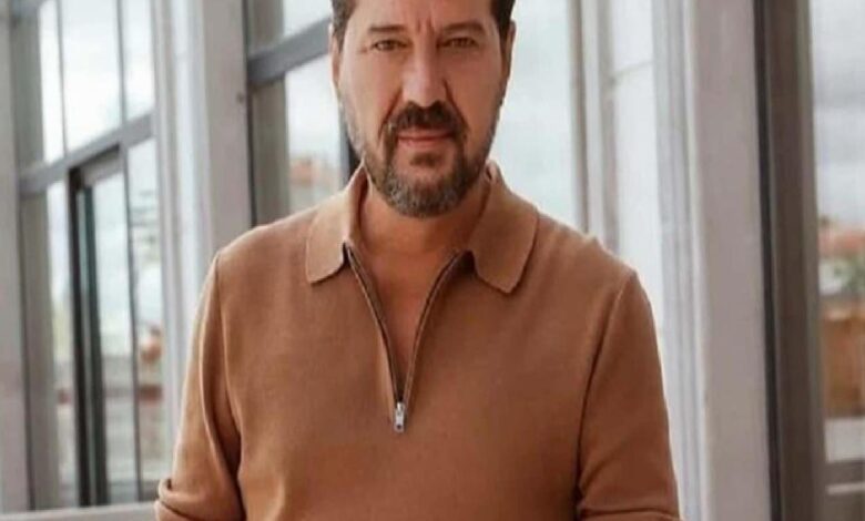 Man with short dark hair and beard wearing a tan zip-neck sweater stands in a glass-walled corridor or atrium, looking toward the camera.