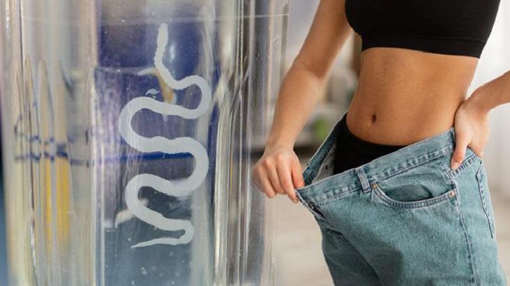 Woman in a crop top holds out baggy jeans at the waist to show weight loss near a water cooler.
