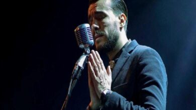 Male singer with hands pressed together in prayer at a microphone on stage.