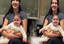 Woman in a gray shirt sits in a kitchen, holding a smiling baby in a white onesie; two mirrored frames visible nearby.