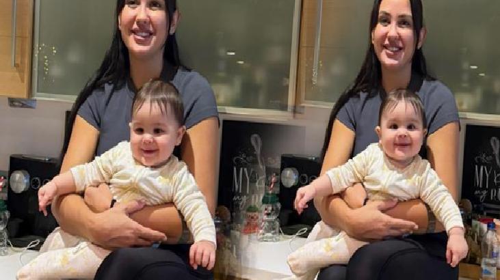 Woman in a gray shirt sits in a kitchen, holding a smiling baby in a white onesie; two mirrored frames visible nearby.